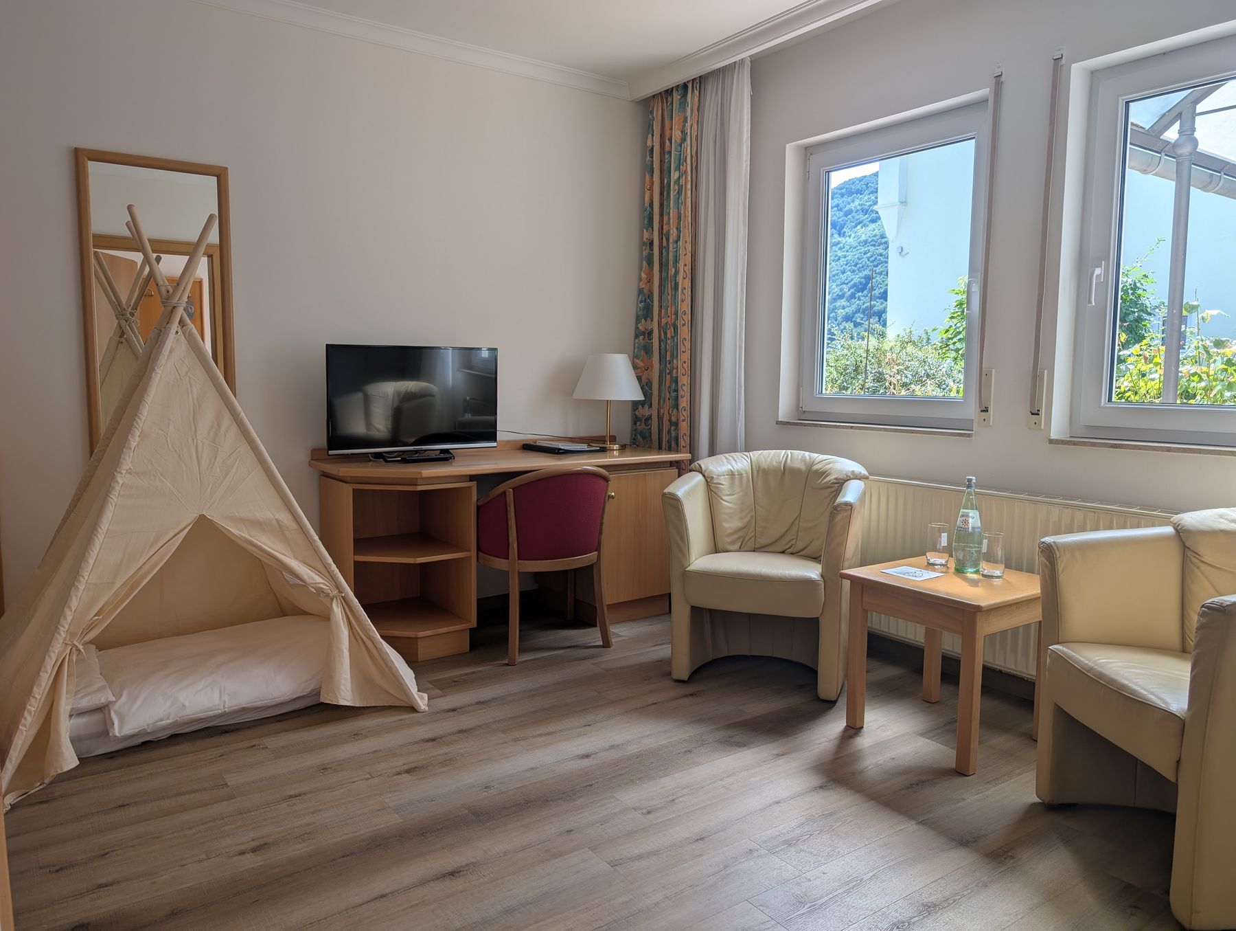 Großes Doppelzimmer mit zwei Sesseln, Schreibtisch, Fernseher und zwei Fenstern, dazu ein Spielzelt für kleine Gäste