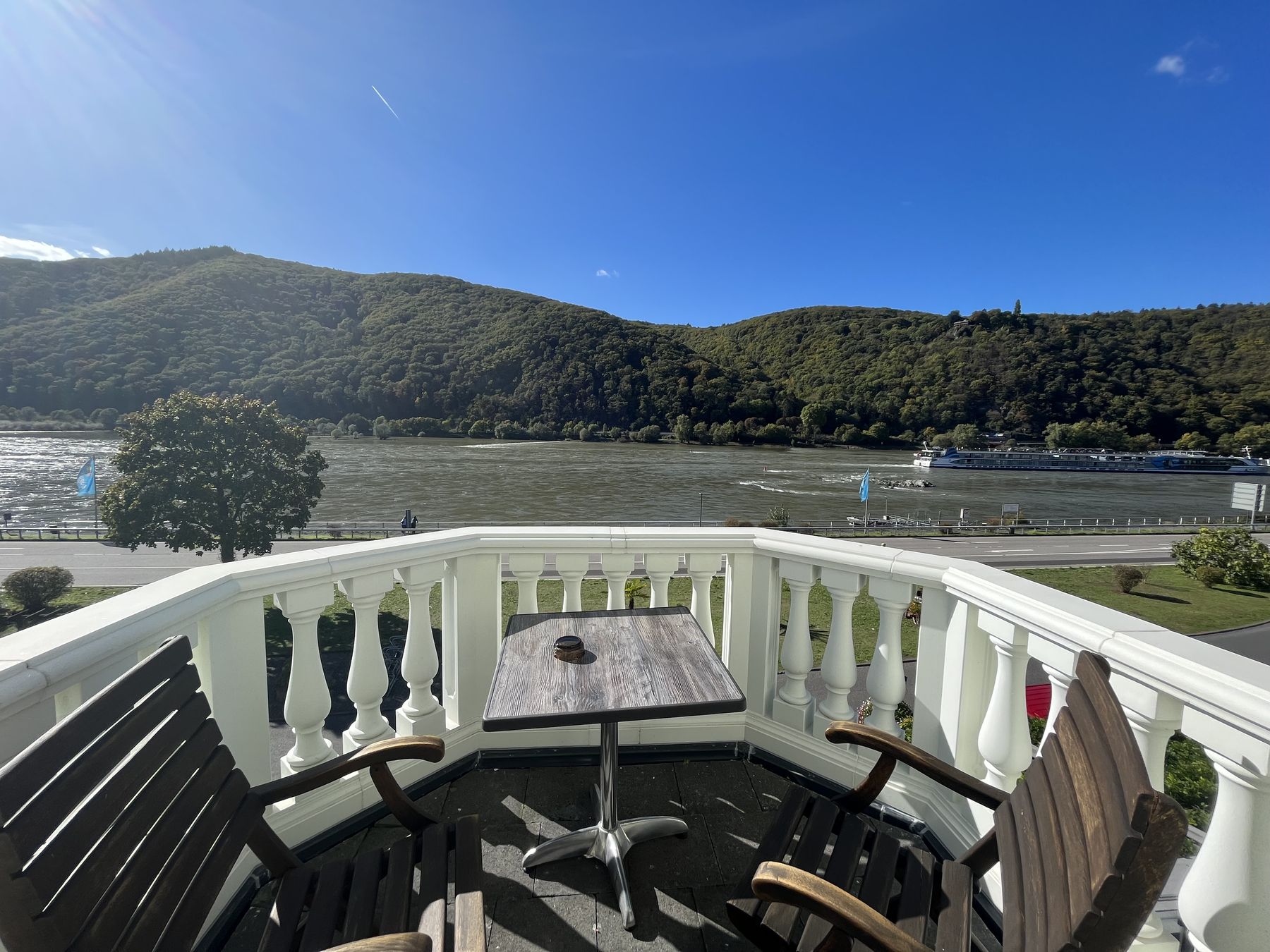 Balkon der Junior Suite mit weißer Balustrade, Holzstühlen und Tisch, davor der Rhein und der bewaldete Hang
